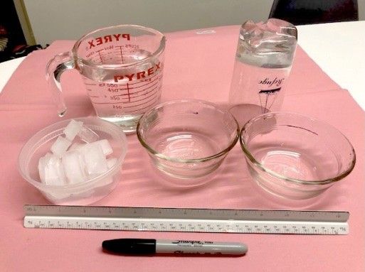 Lab setup with bowls, ice cubes, measuring cup of water, ruler, and marker on a pink surface.