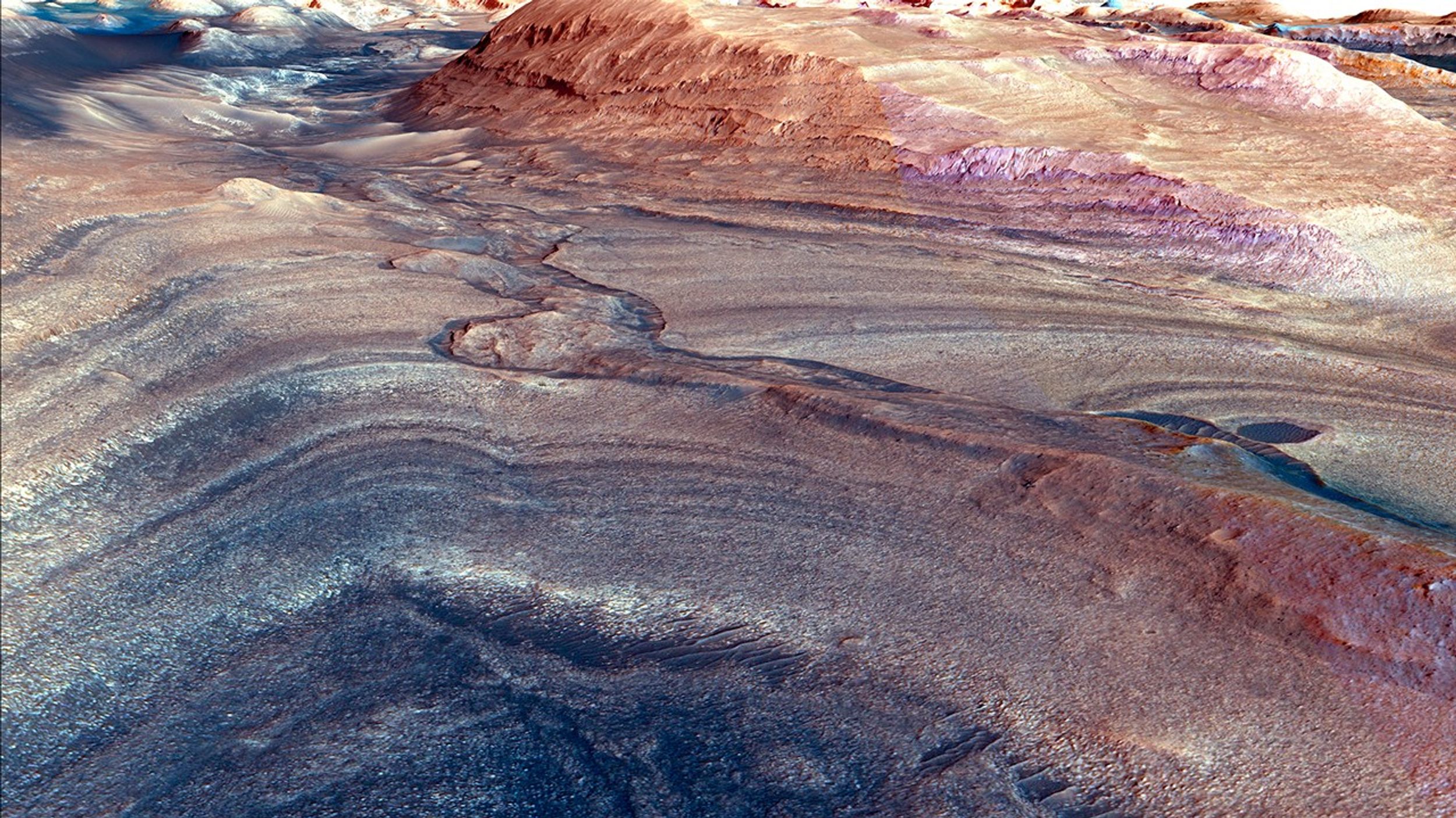 Visualización obtenida con el Orbitador de Reconocimiento de Marte (MRO) de la NASA que muestra el diverso paisaje geológico del cráter Gale de Marte, que el rover Curiosity de la NASA ha estado explorando. La imagen captura una pendiente pronunciada con distintas formaciones rocosas en capas de diversos colores, incluyendo tonos de marrón rojizo, marrón claro y morado. La parte superior de la imagen muestra el canal Gediz Vallis, mientras que más abajo son visibles franjas de depósitos sedimentarios creados a lo largo de millones de años. Estas capas contienen sulfatos y otros minerales que se formaron a medida que el agua se evaporaba, lo que aporta evidencia del antiguo pasado acuoso de Marte. Las abundantes variaciones geológicas del terreno de Marte ofrecen información vital sobre la historia de su clima y la posibilidad de que haya sido habitado por vida microbiana hace unos 3.700 millones de años. Esta imagen en color realzado fue creada en marzo de 2024 con datos de la cámara HiRISE y otros instrumentos a bordo de MRO.