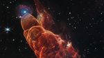 A stunning James Webb Space Telescope image showing Herbig-Haro 49/50, nicknamed the 'Cosmic Tornado.' The foreground features dramatic reddish-orange swirls and wisps of gas, representing outflow from a young forming star, stretching across the dark background of space. In the upper left, a blue-centered spiral galaxy is visible behind the outflow, with its central bulge glowing blue and reddish star-forming regions in its spiral arms. Numerous stars dot the black cosmic background. This detailed infrared observation reveals the processes of star formation and jet activity that help scientists understand the origins of our own solar system.