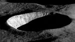 A black and white photograph of an unnamed lunar crater captured by NASA's Lunar Reconnaissance Orbiter Camera (LROC) on August 30, 2023. The image shows dramatic contrast between light and shadow as early morning sunlight illuminates only the western wall of the crater, which appears bright white against the dark lunar landscape. The interior of the crater remains in complete shadow, creating a striking bowl-shaped void. The surrounding lunar surface displays the characteristic rough, textured terrain of the Moon with various small craters and ridges visible. This high-resolution image demonstrates LROC's capability to capture detailed topographical features, part of LRO's mission to map the lunar surface for future exploration efforts.