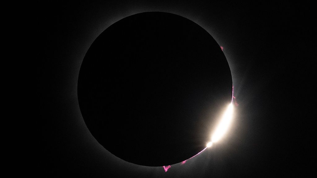 A dramatic photograph of the April 8, 2024, total solar eclipse taken from Dallas, Texas, showing the moment just after totality when the Moon blocks most of the Sun's disk against a black background. The Sun's outer atmosphere (corona) is visible as a faint white glow surrounding the Moon's silhouette. Several bright pink solar prominences—jets of plasma extending from the Sun's surface—are clearly visible along the edges of the Moon, particularly pronounced at the bottom and right edges. The lower right portion of the image shows "Bailey's beads," appearing as bright white segments where sunlight streams through valleys on the lunar limb, creating a diamond ring effect. This rare astronomical phenomenon allowed scientists to study the innermost part of the corona that is only observable during total eclipses, providing valuable data about solar energy transfer mechanisms.