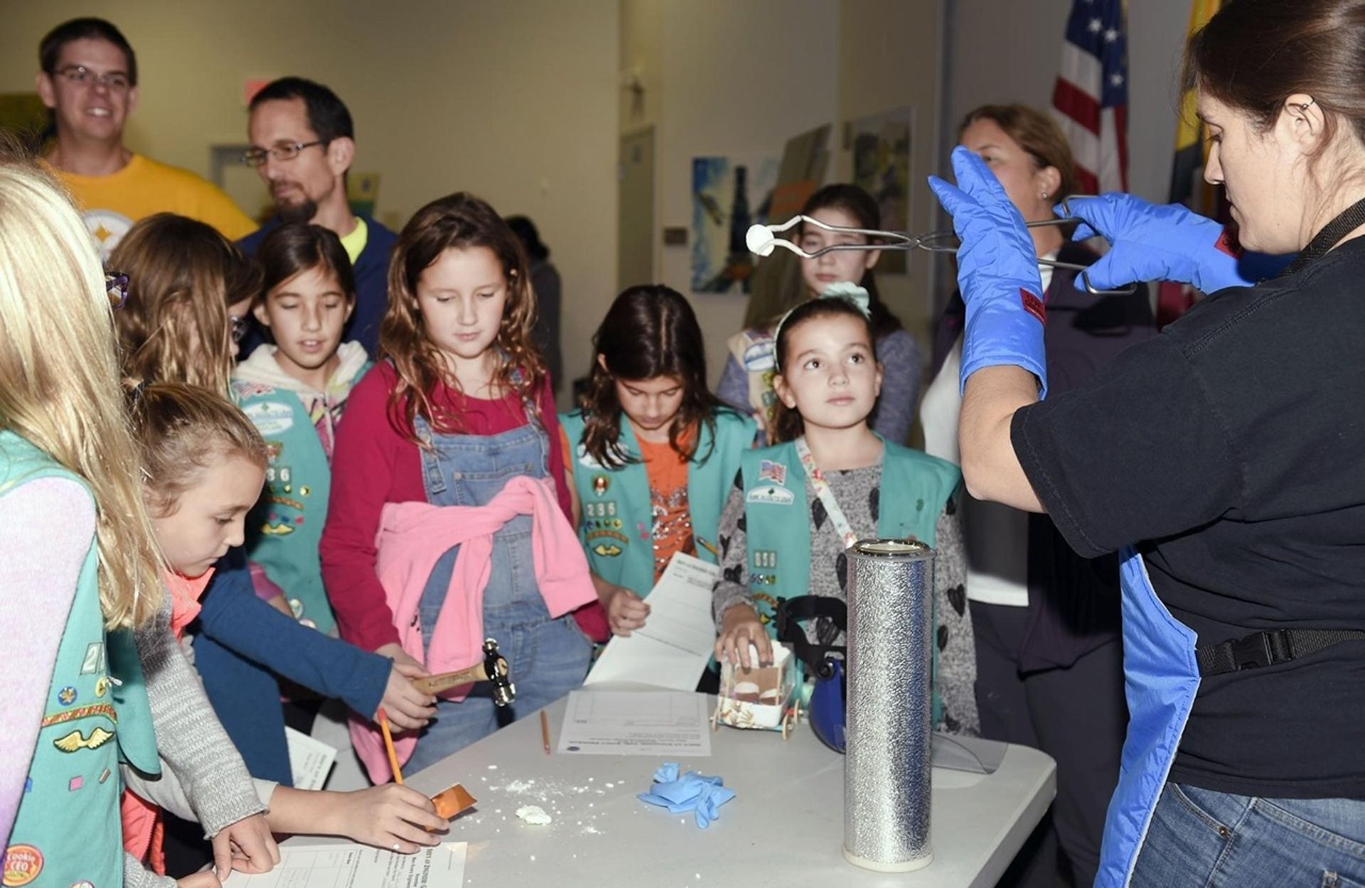 A group of Girl Scouts gathers around a table during a science demonstration. A woman wearing blue gloves uses tongs to handle a marshmallow over a container, likely involving liquid nitrogen, while the children observe and take notes.