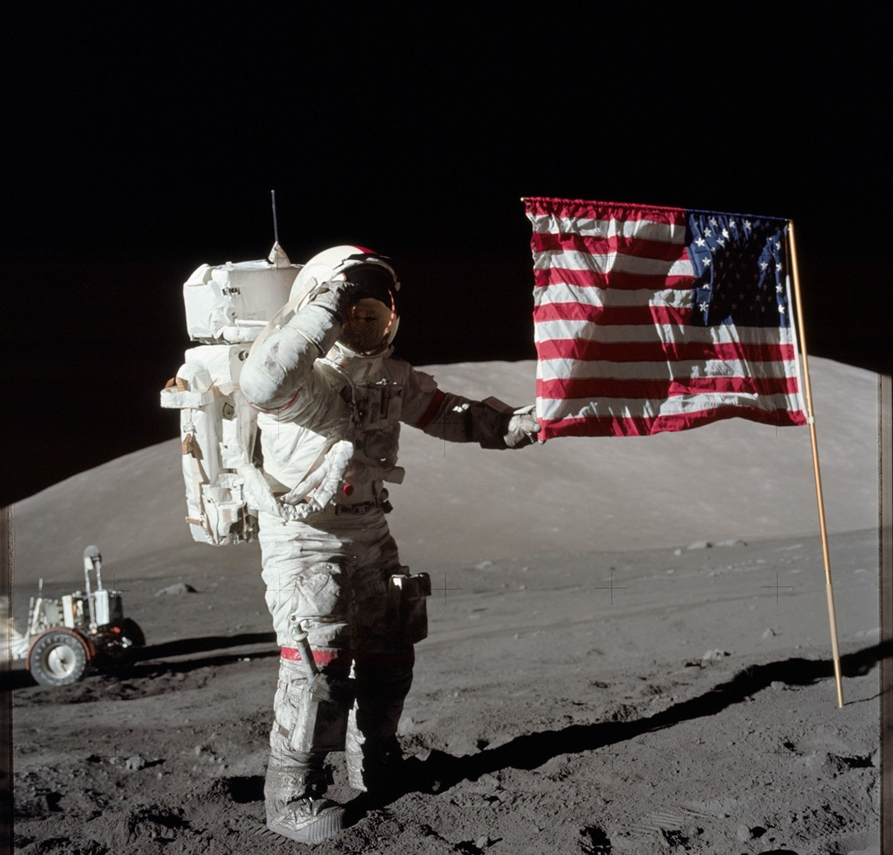 Astronaut in a white space suit salutes while standing on the Moon next to an American flag. Lunar rover visible in the background, with a barren, dusty lunar surface under a black sky.