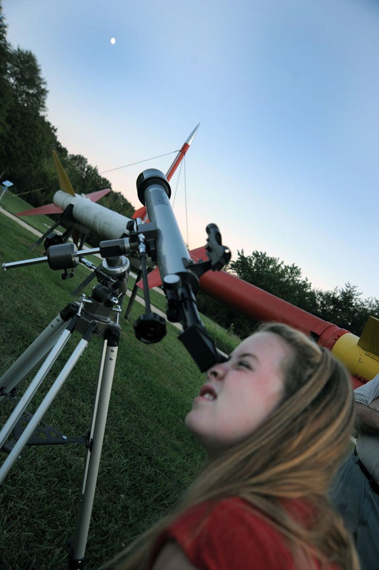 A young girl looks through a telescope at the Moon during twilight, with a large outdoor rocket display in the background.