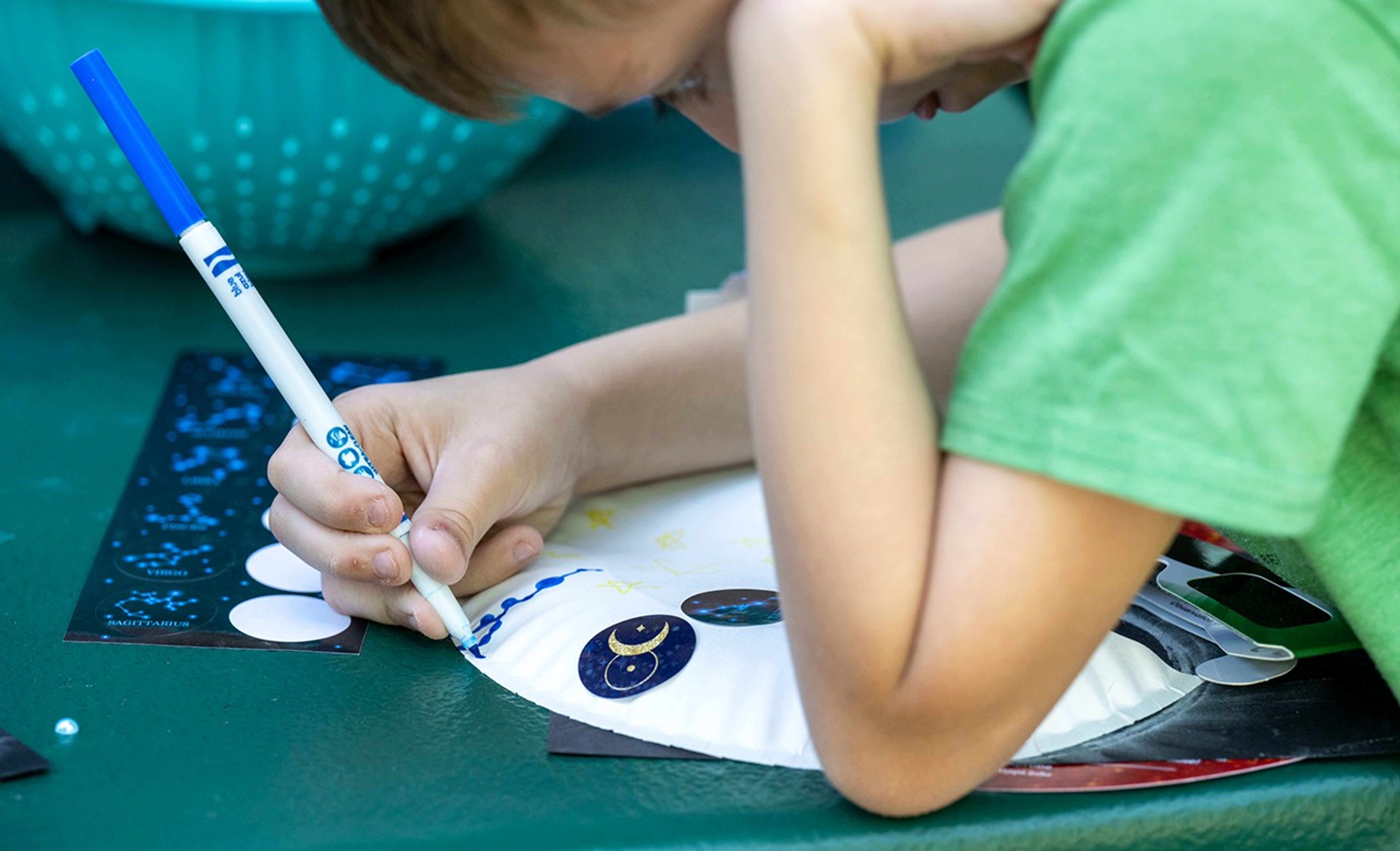 A child in a green T-shirt colors in the back of a paper plate with a blue magic marker.