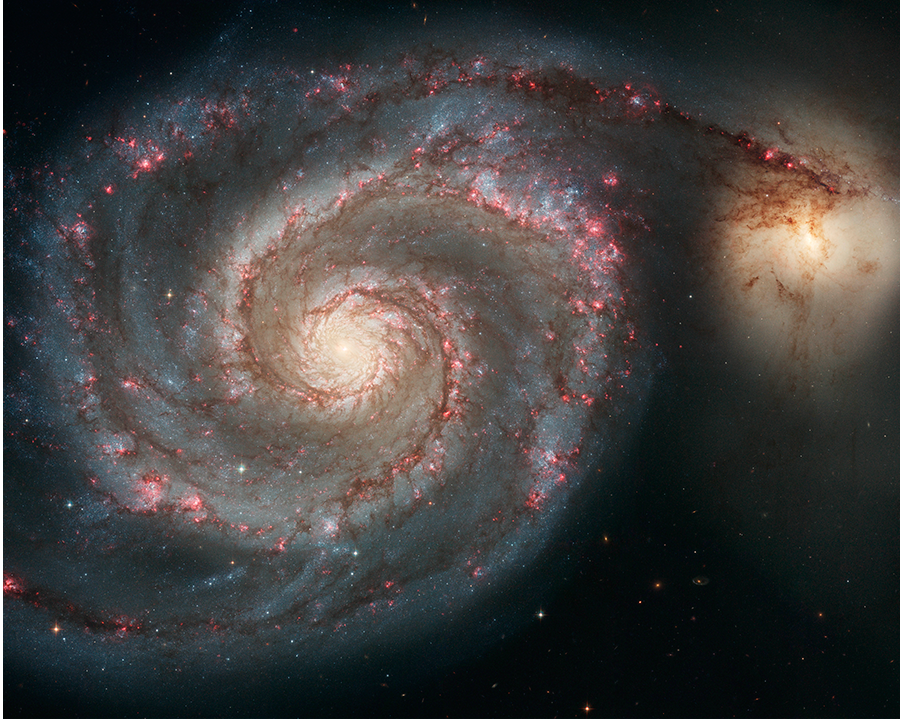 Una gran galaxia espiral con brazos distintivos llenos de franjas de polvo de color marrón rojizo y regiones de formación estelar de color rojo rosáceo. Uno de los brazos espirales se extiende hacia la derecha y, al final de este brazo, hay otra galaxia. Parece una brillante bola de estrellas de color blanco amarillento.