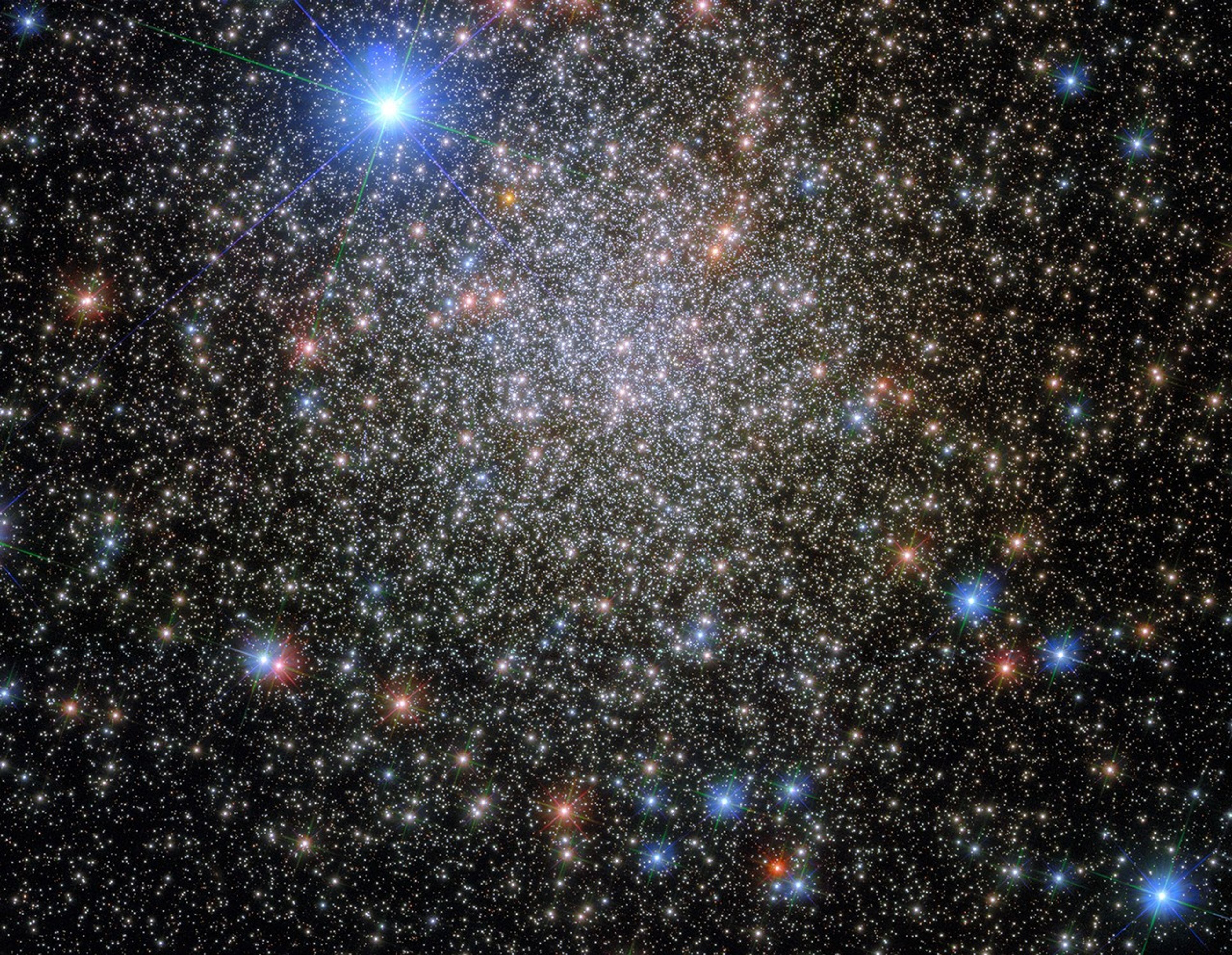 A thick group of many thousands of whitish stars as the center. Near the edges, where the stars are thinner, some blue and red-orange stars are visible. A very bright blue-white star shines in the foreground in the upper-left.