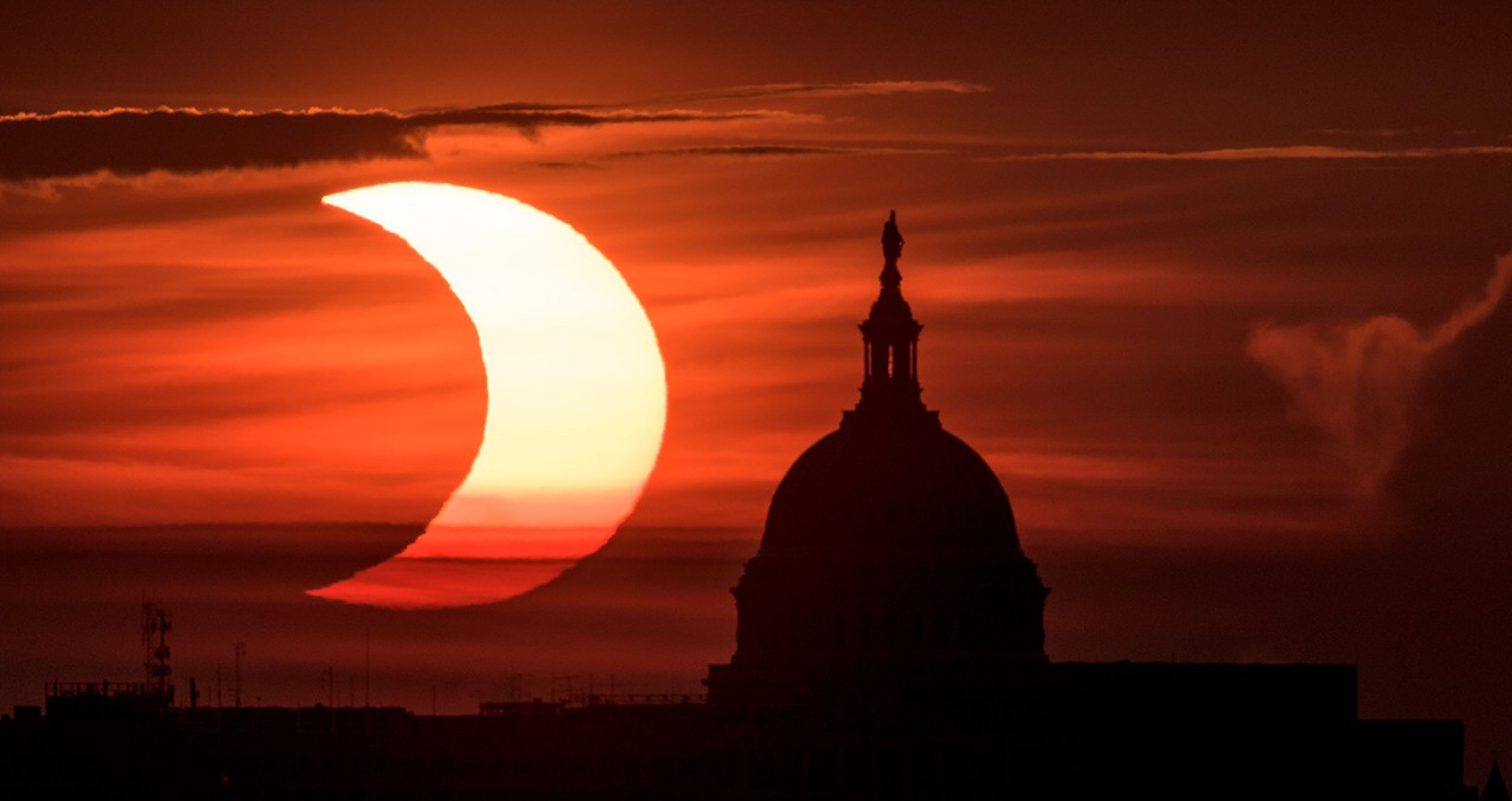 Un Sol en forma de media luna sobre un fondo rojo y naranja oscuro. Está a la izquierda de la parte superior del edificio del Capitolio de los Estados Unidos, que se muestra en silueta.
