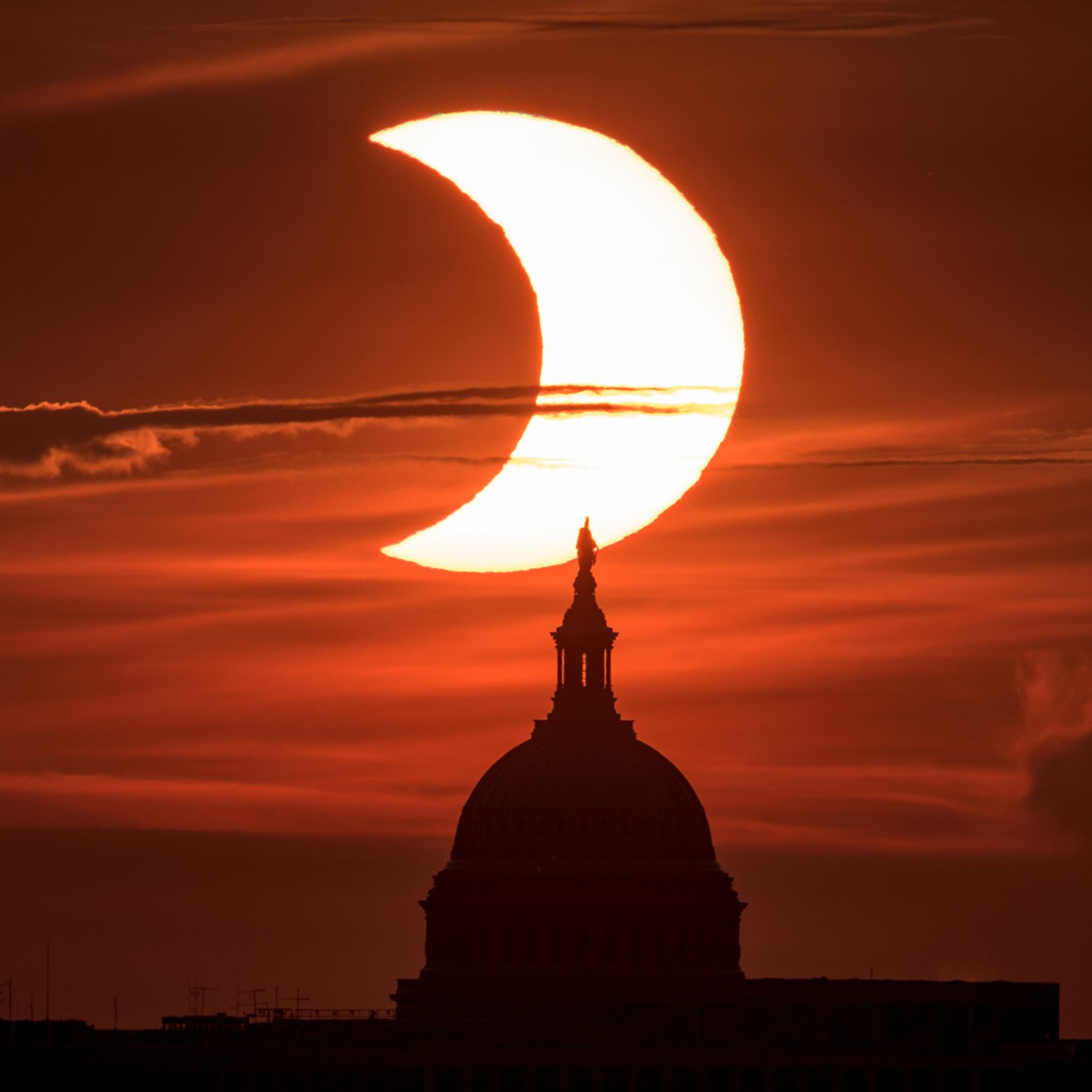 A crescent Sun against a dark orange and red background. It's behind the top of the U.S. Capitol Building, shown in silhouette.