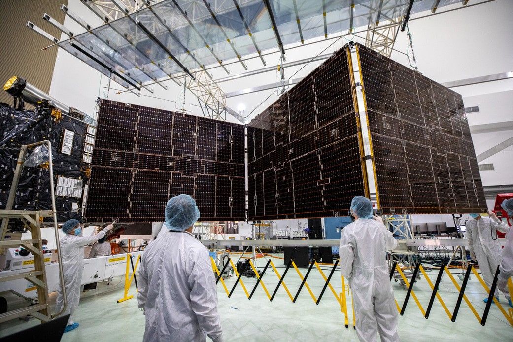 Workers in white protective clothing, blue hair covers, and face masks watch as the solar arrays for Psyche are retracted.