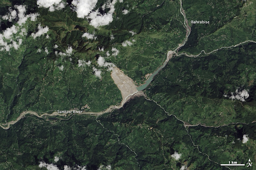 A true-color satellite image shows a green, mountainous area bisected from top right to bottom left by a large river. Clouds float over the peaks. A large brown-gray triangular scar starts at the top of a mountain and widens down to and across the riverbed. The river has cut through this material and washed it downstream, as evident by the brown color in the riverbed, which contrasts with the green color of the river upstream of the slide.