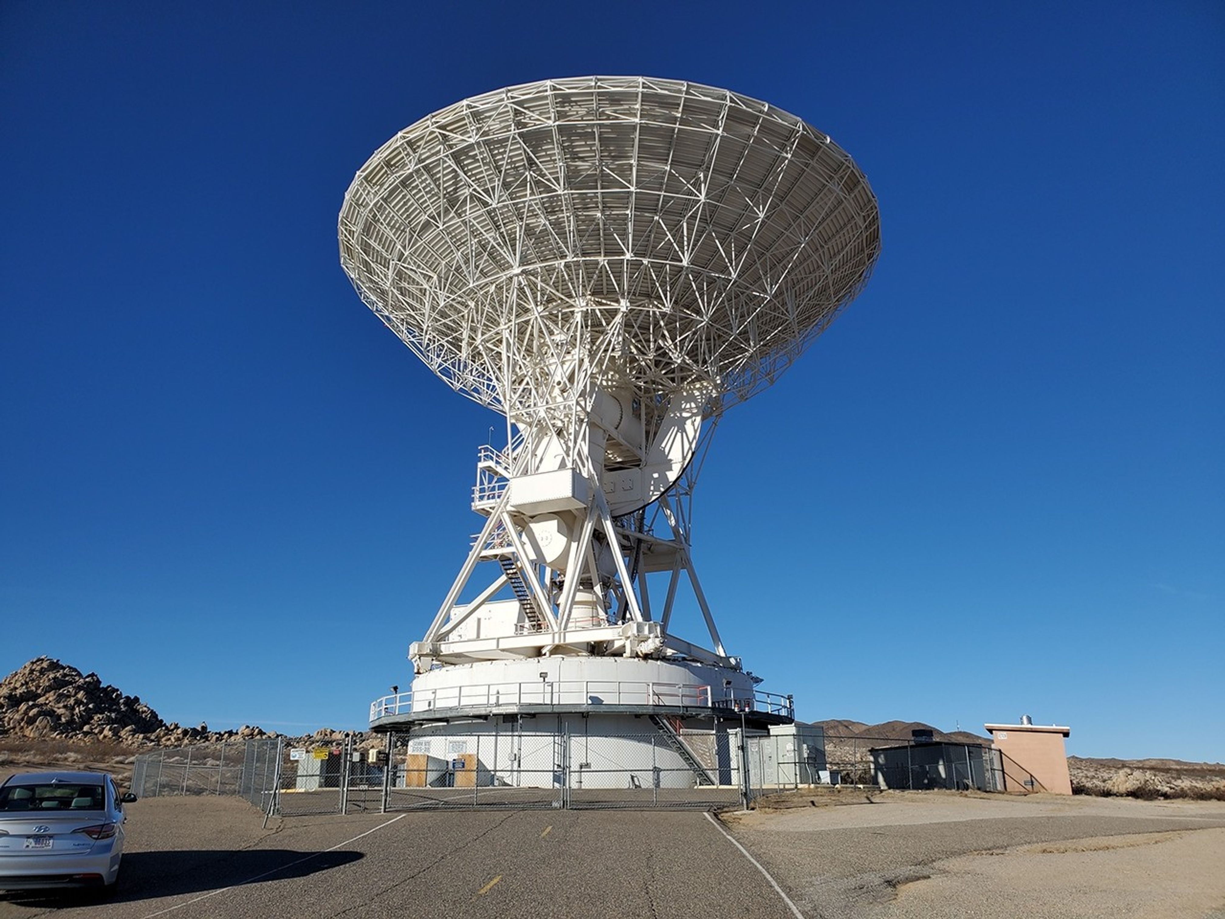 Against a deep blue sky and on a concrete ground, a large white disk faces upward. There are stairs leading from the base to the dish itself.
