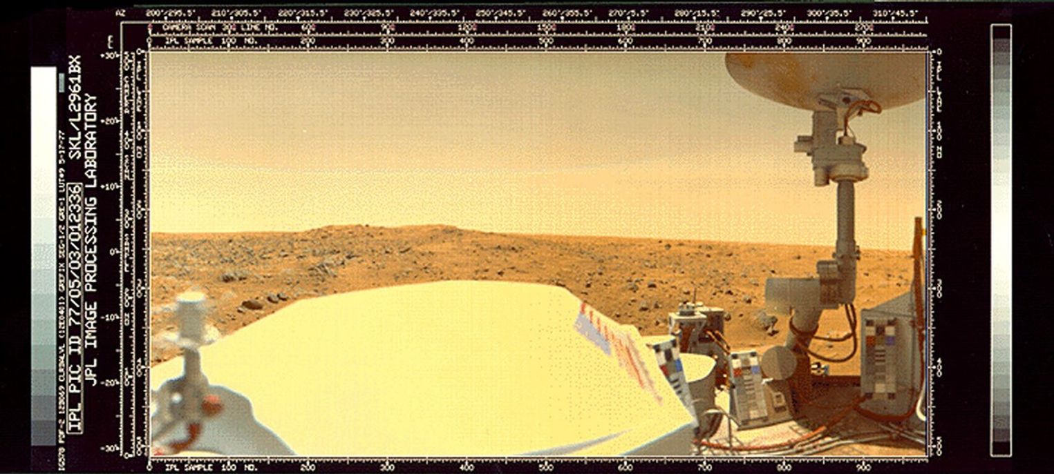 A rocky plain on Mars is visible over the top of the Viking 1 lander. Part of the top deck of the lander, including a large antenna dish, are visible in the foreground.