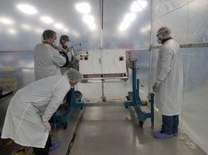 Four technicians in white cleanroom suits, hairnets, and glasses are working around a rectangular white scientific instrument mounted on a blue wheeled cart. The instrument has several connectors and panels visible on its side. One technician is leaning over the front of the cart, another is positioned behind the instrument appearing to make adjustments, and a third is standing to the far right, observing. The fourth technician is partially visible behind the instrument. They are working inside what appears to be a temporary cleanroom enclosure with translucent plastic walls and bright overhead lighting.