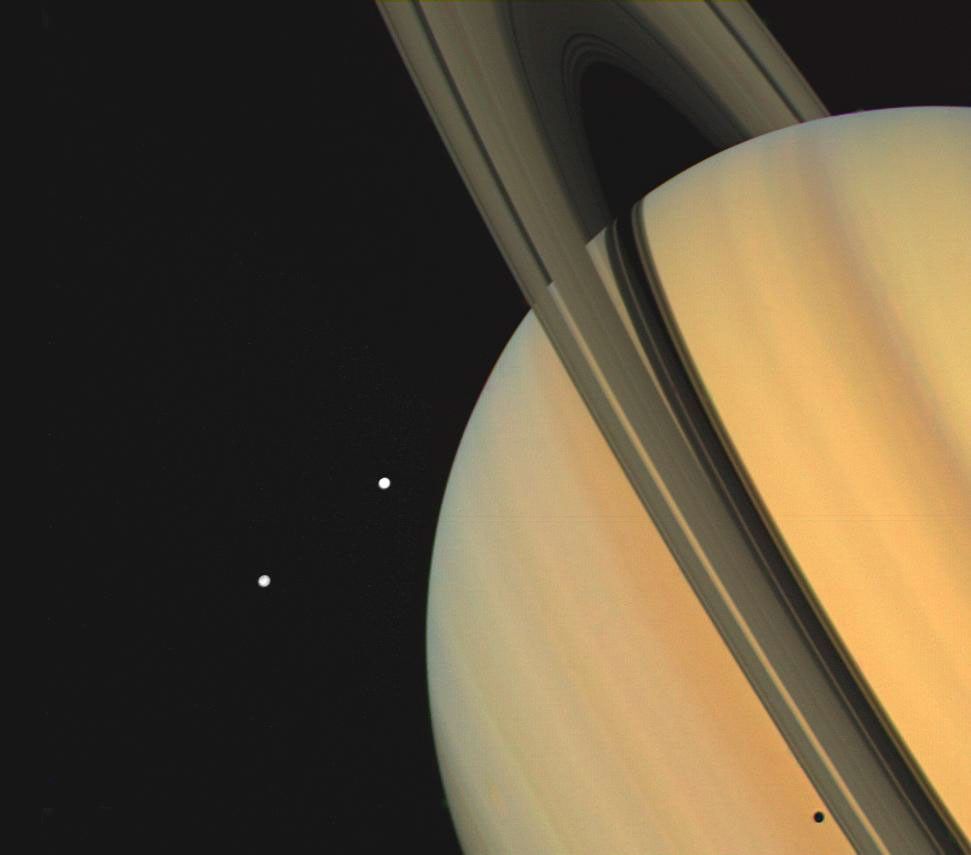 Saturn appears golden with darker gold and reddish-brown bands. Part of the planet's ring system is visible making the planet appear tilted. Two bright white dots are off the left side of the image. The dots are two of Saturn's dozens of moons.