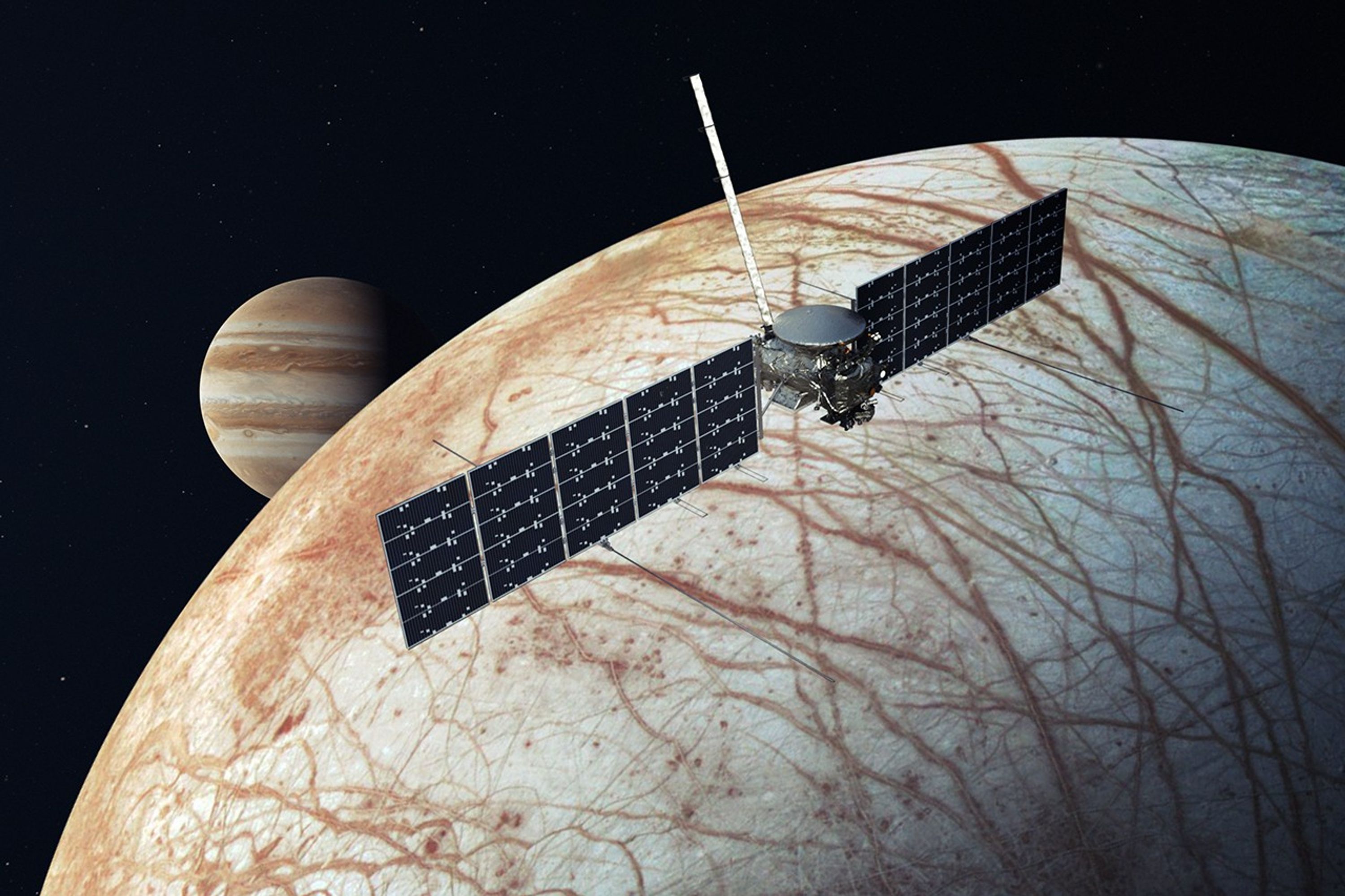 Artist's rendering of NASA's Europa Clipper spacecraft, shown as a small metallic gray and silver body with two very long, black solar arrays extending from either side., as it flies over Europa. The moon fills most of the image, with its surface shades or grayish white and light tan, crisscrossed throughout with rust-colored veins. Jupiter is visible in the background at upper left, slightly obscured by Europa, recognizable by its alternating horizontal cloud bands, in shades of orange and gray, against a black backdrop of outer space.
