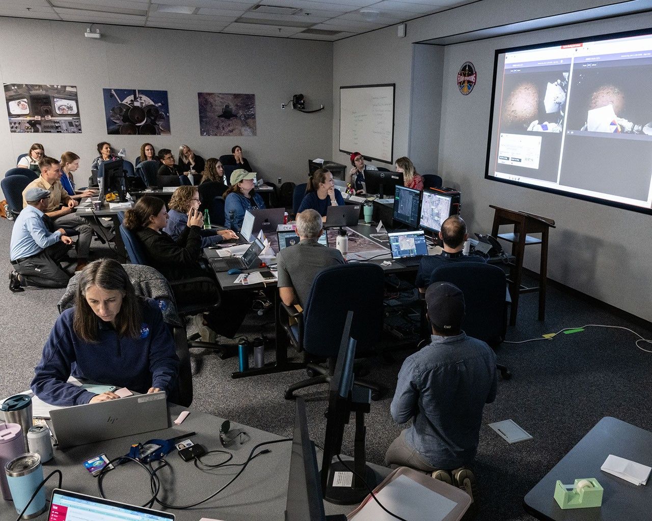 People are sitting in a typical, brightly lit office-building room. Colorful posters line the walls. A large screen is in the top right corner, showing two side-by-side images of shapes that are hard to make out. People are sitting around tables, some are kneeling, looking either at the large screen or at the small computer screens in front of them.