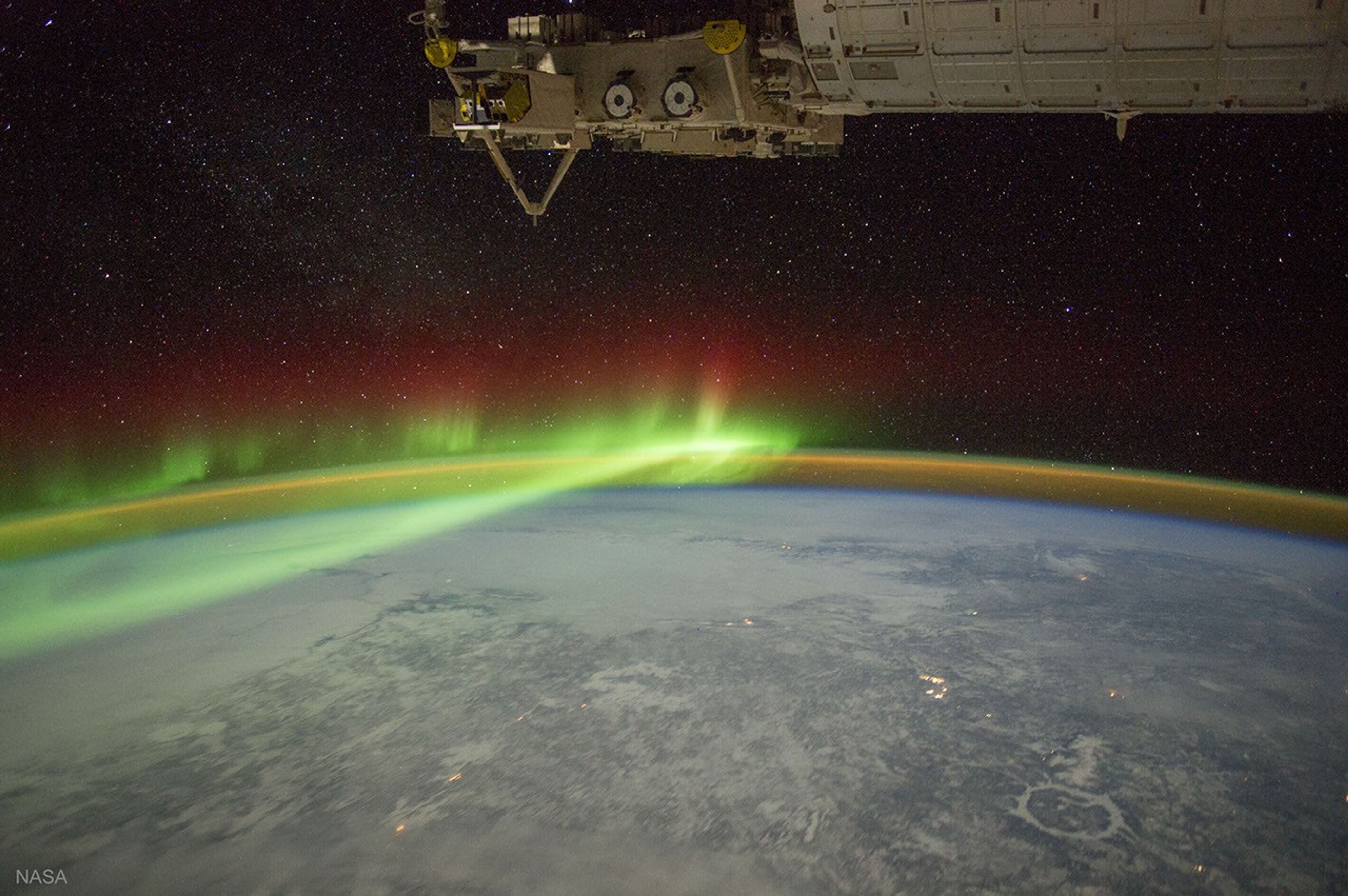Image of Earth at night with lights glowing on Earth�s surface. Above Earth are green, and red bands of light signifying airglow. Above the bands of light is a black space with white stars and an orange and green band of light below the airglow.