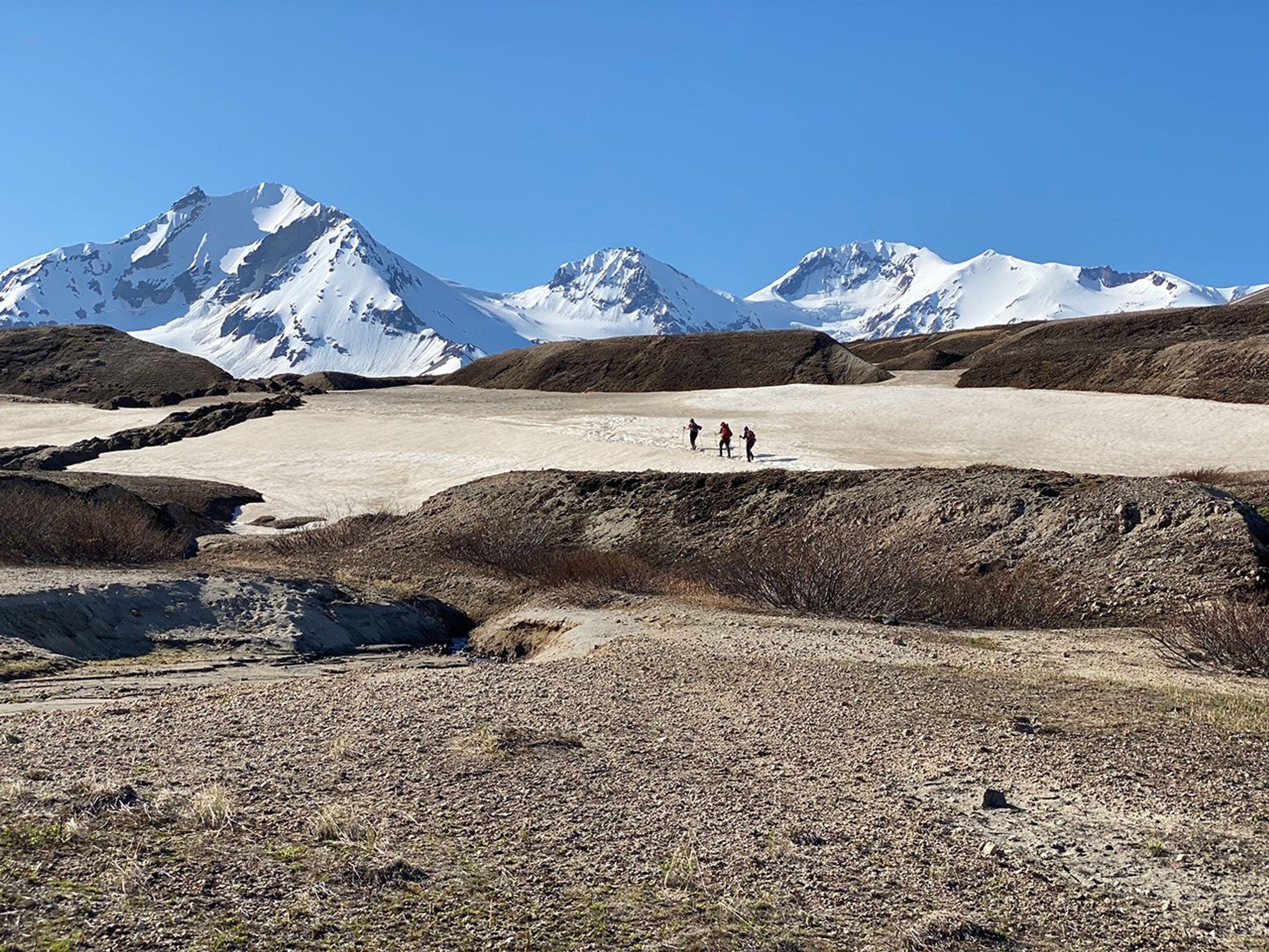 Three people, wearing large backpacks, trek across a snow field between hills of dark rubble. In the background: steep, snow-covered mountains under a blue sky.