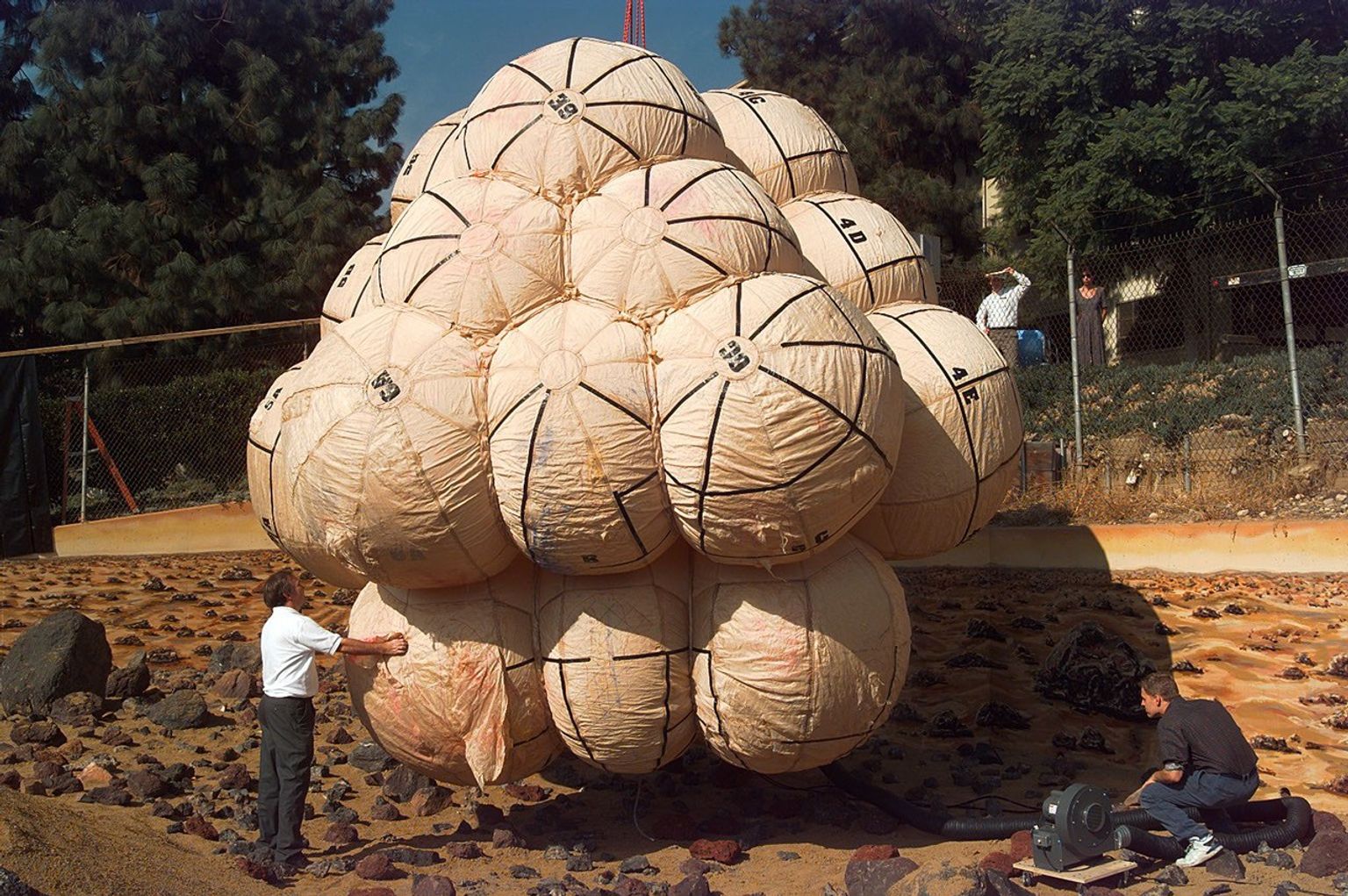 A man dressed in dark slacks and a white shirt stands outdoors at image left, examining a giant inflated airbag composed of numerous large, fabric, roughly spherical balloons, each almost as large as the man, all attached to each other, with more than a dozen visible. Another man in blue jeans and black shirt crouches at lower right, operating what appears to be an air blower, which has a wide black hose extending to the underside of the airbag package.