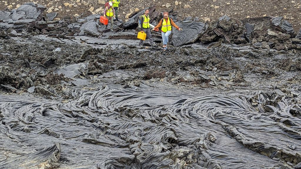Scientists wearing yellow-green reflective vests and carrying equipment cases carefully step across a dark gray lava flow.