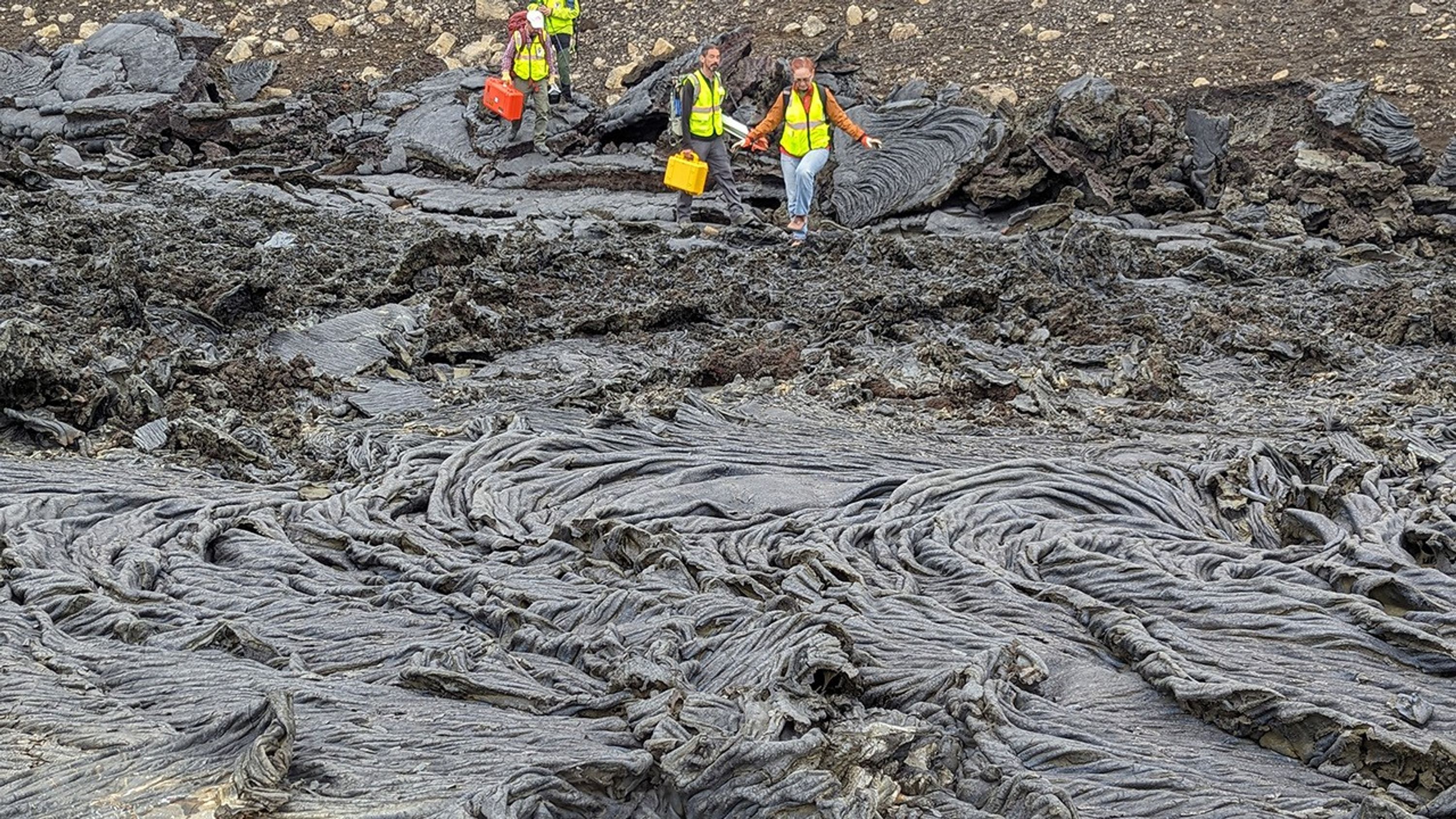 Scientists wearing yellow-green reflective vests and carrying equipment cases carefully step across a dark gray lava flow.