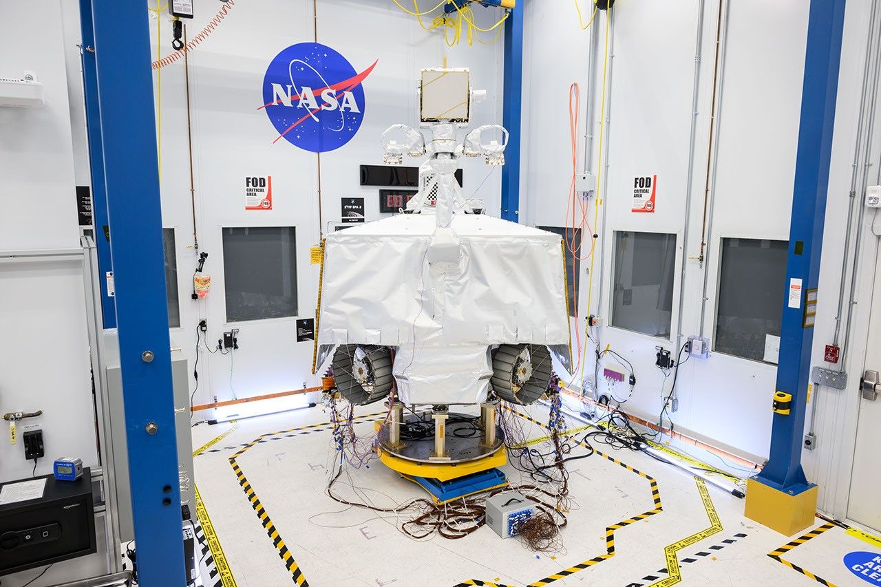 NASA's VIPER robotic Moon rover is shown in a clean room at NASA's Johnson Space Center in Houston. Two of its wheels are visible in this image. It is shown on a white floor in front of a white wall next to the NASA "meatball" logo. On the the floor there is yellow-and-black tape surrounding the rover.