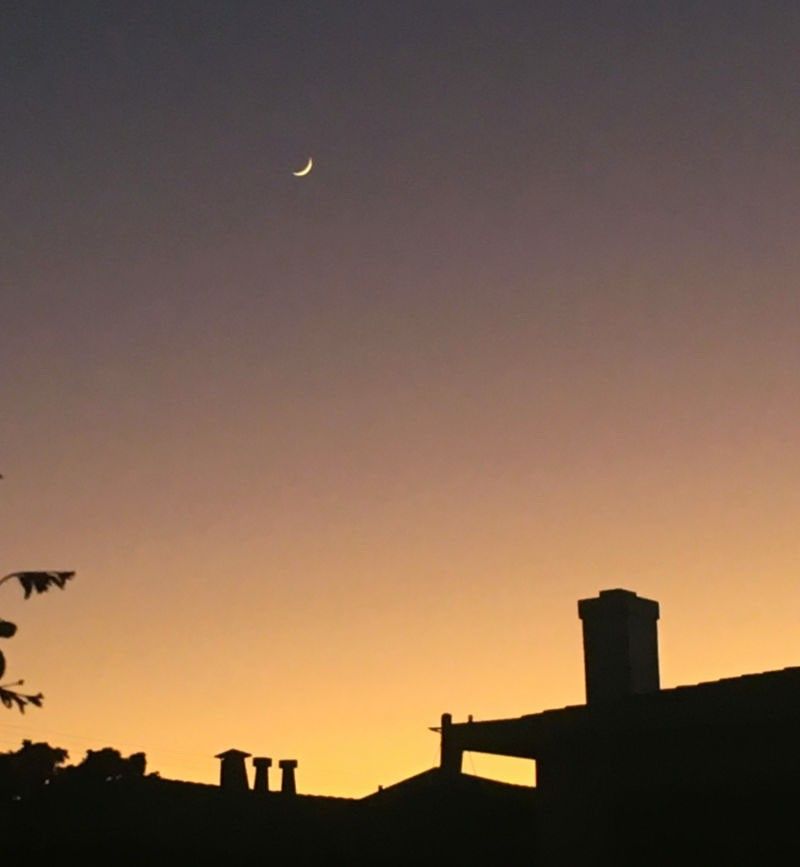 Foto de una Luna creciente suspendida sobre la silueta de un tejado al atardecer.