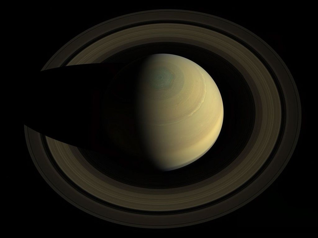 This image looks down on Saturn. The planet and its rings appear to have muted shades of gold and brown.  The planet is casting a shadow on its rings on the left side of the image.