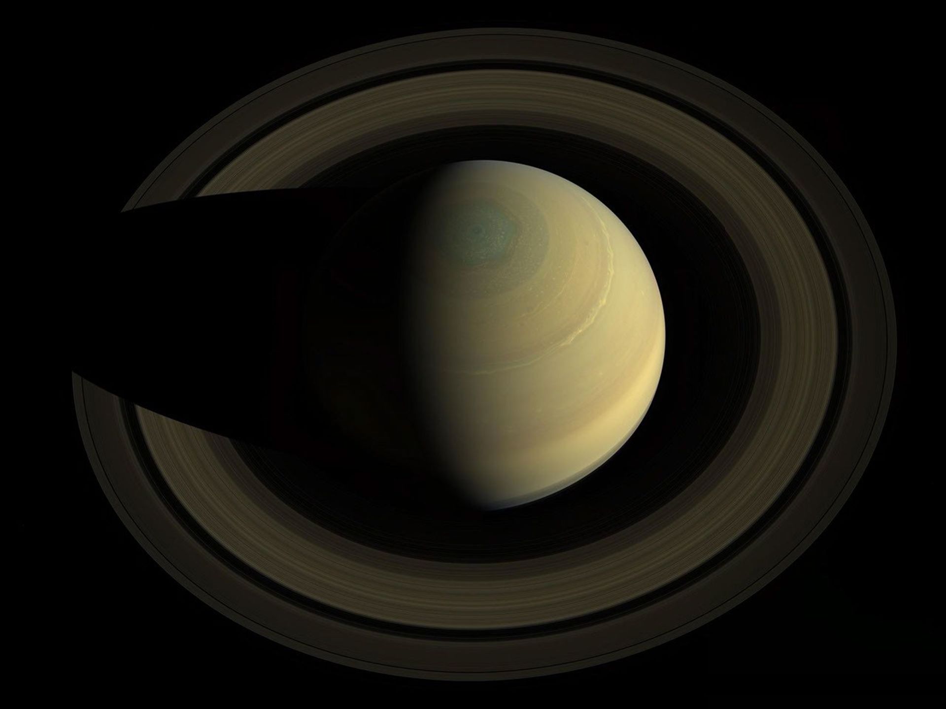 This image looks down on Saturn. The planet and its rings appear to have muted shades of gold and brown. The planet is casting a shadow on its rings on the left side of the image.