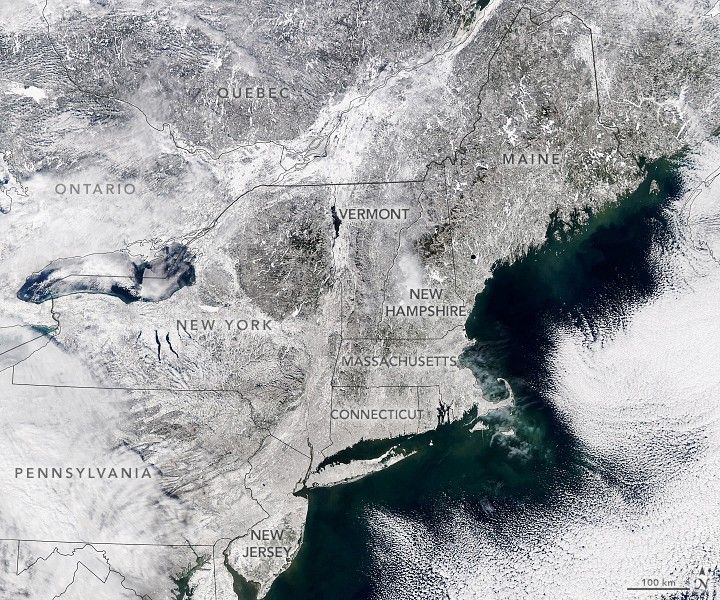 The landmass of the northeastern U.S. and parts of eastern Canada are covered in white from snow. The outline of the states of the northeastern U.S. and providences in eastern Canada are in black. The states are labeled in black and are listed from bottom to top as Pennsylvania, New Jersey, New York, Connecticut, Massachusetts, Vermont, New Hampshire, and Maine. The state of Rhode Island is also shown, but not labeled. The eastern Canadian providences of Ontario and Quebec are also labeled in black and are located above New York and to the left of Maine. There is dark green-blue water next to the northeastern U.S. coastline. There are white clouds to the right of the water, covering some of the water.