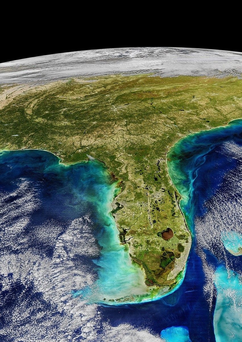 The image is of Florida and the southeastern U.S. from space. The land is green and tans and the ocean is different shades of blue. Along the coast of Florida’s peninsula is a bright turquoise color. A large lake in Florida is brown. There are white clouds over the land to the north and in the ocean around the peninsula. The very top of the image is the black of space.