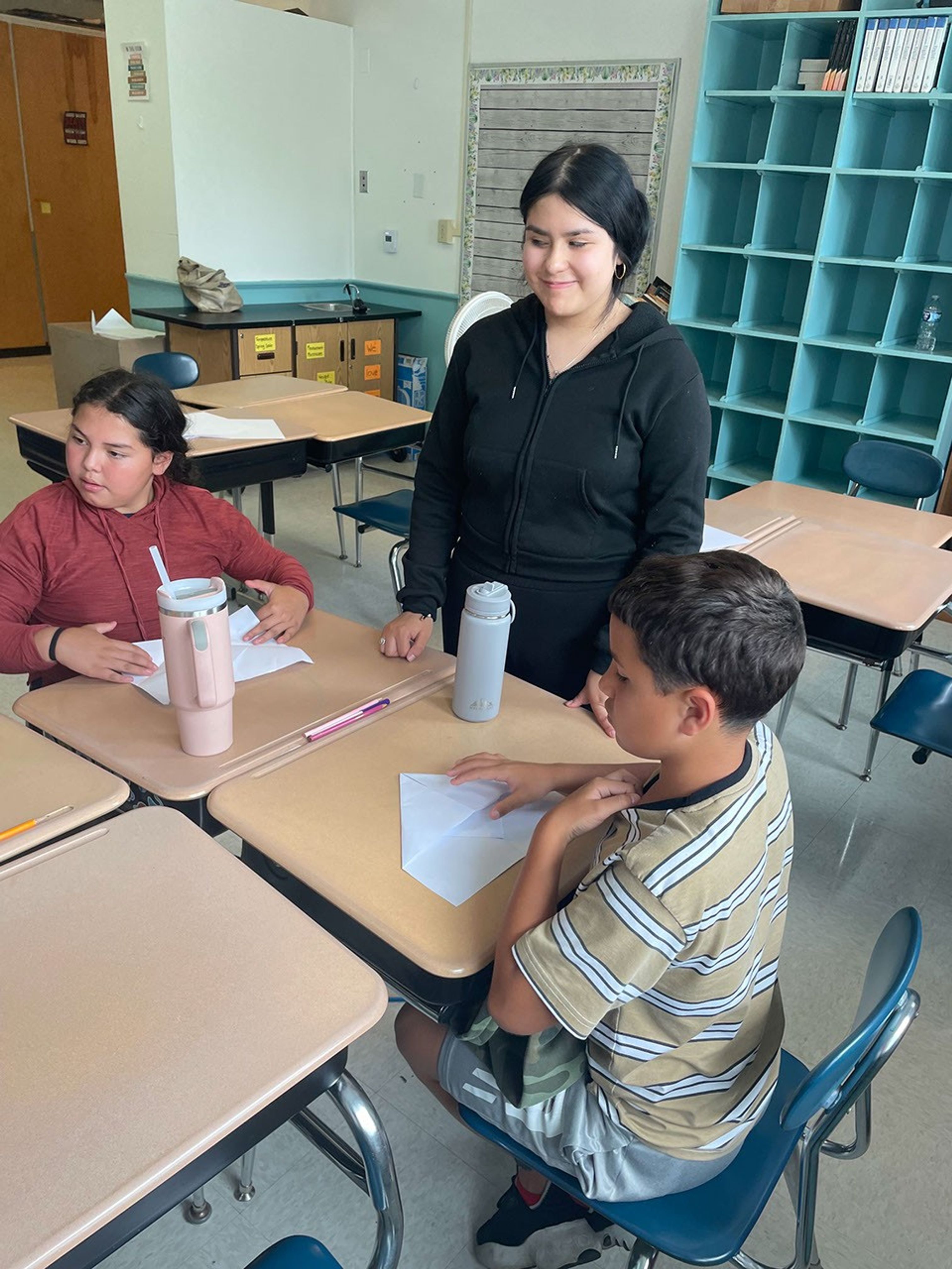 Two youth sitting at desks with paper in front of them with an adult educator standing between them.
