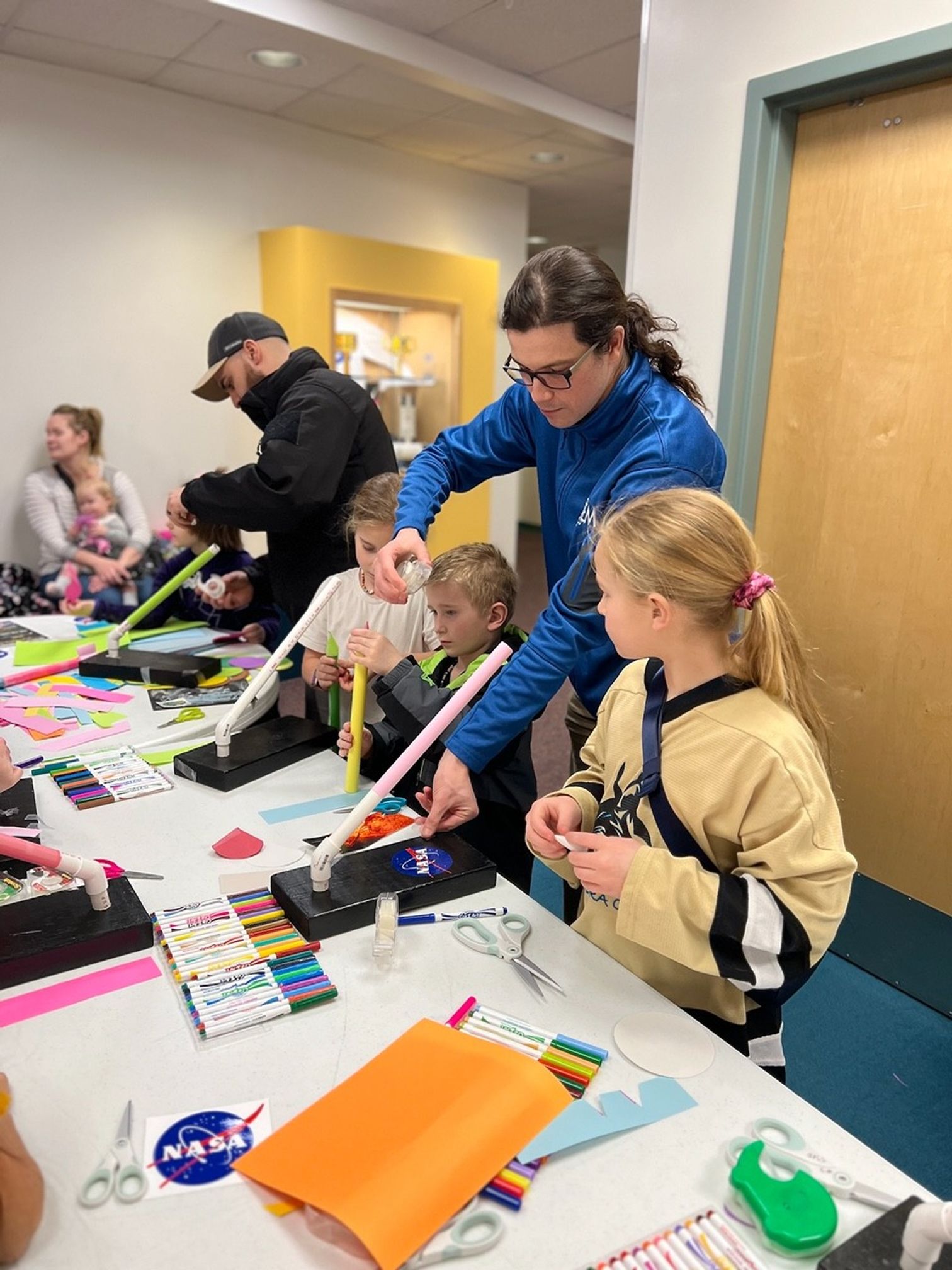 Several children were on one side of the table that had rolled up papers, color markers, and stickers. Two adults were helping them. A third adult was seated and holding a child.