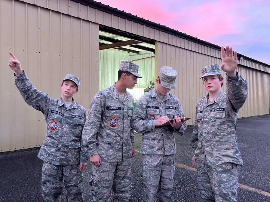 Four youth in Civil Air Patrol uniforms collecting data. Two are pointing at the sky, and a third is making notes on a clipboard, while a fourth looks on. In the background is an aircraft hanger.