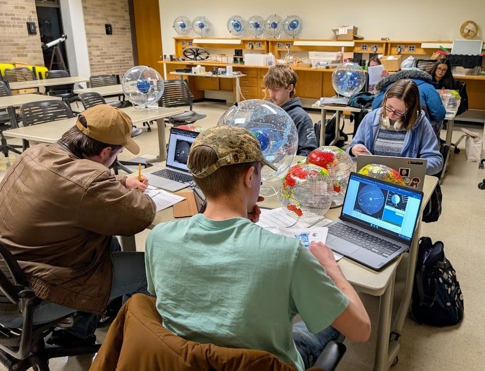 Classroom with seven students sitting at desks. They have their laptops open showing images of the celestial sphere. On the desks between the students are Earth globes and model celestial spheres.