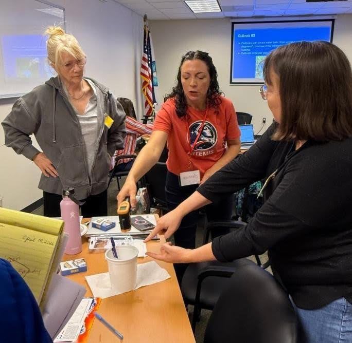 Jessica Taylor demonstrates how to calibrate an infrared thermometer at a table, pointing to materials while two participants watch and follow along in a classroom setting.