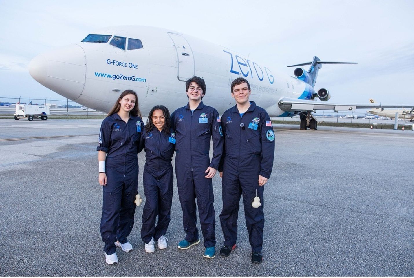 Four young people wearing matching navy blue flight suits stand together, smiling in front of a Zero-G aircraft labeled 'G-FORCE ONE.' The background shows an airport tarmac with other planes and equipment.