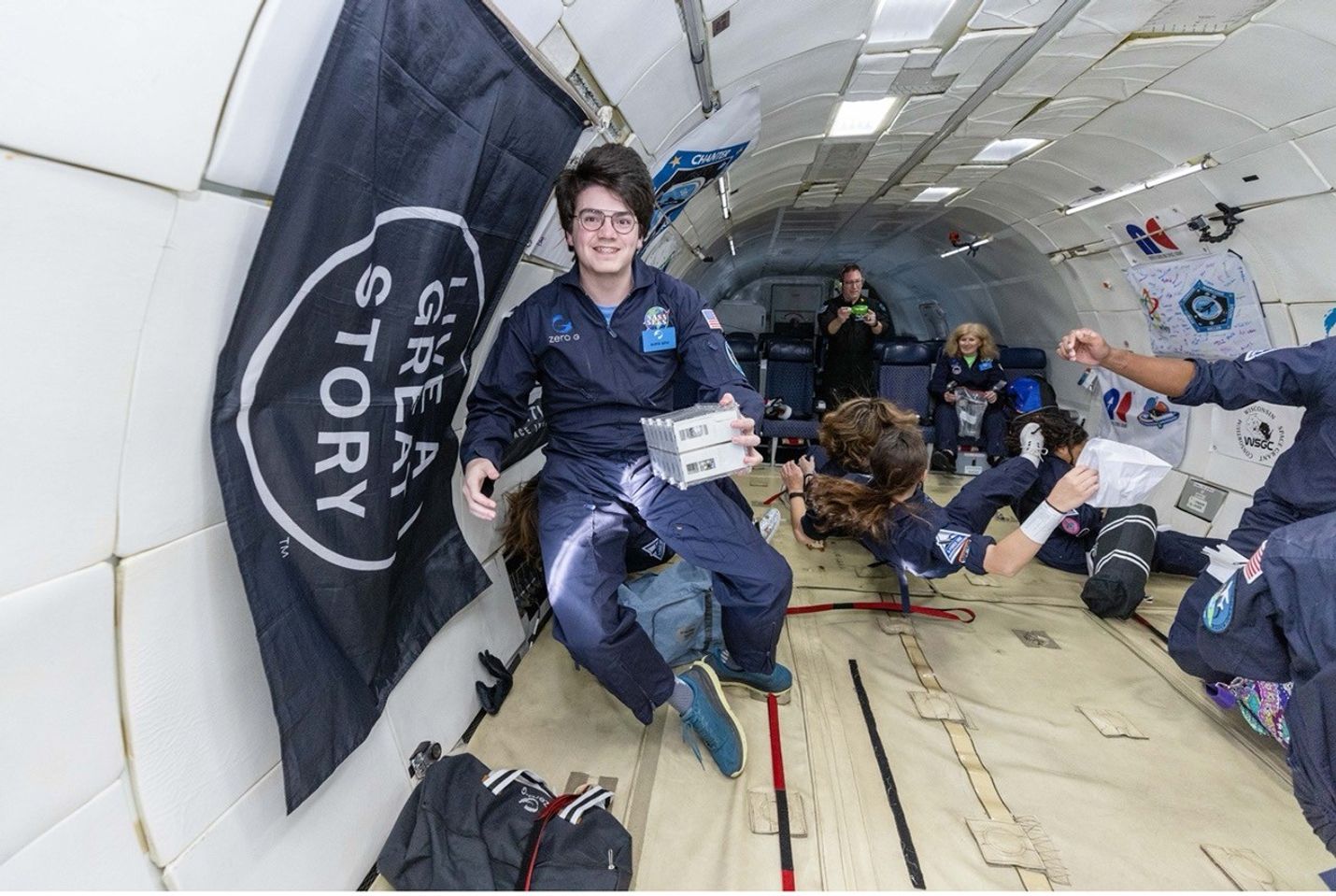 A young person in a navy blue flight suit floats inside a Zero-G aircraft, holding a small experiment module. Other participants are floating in the background, experiencing weightlessness.