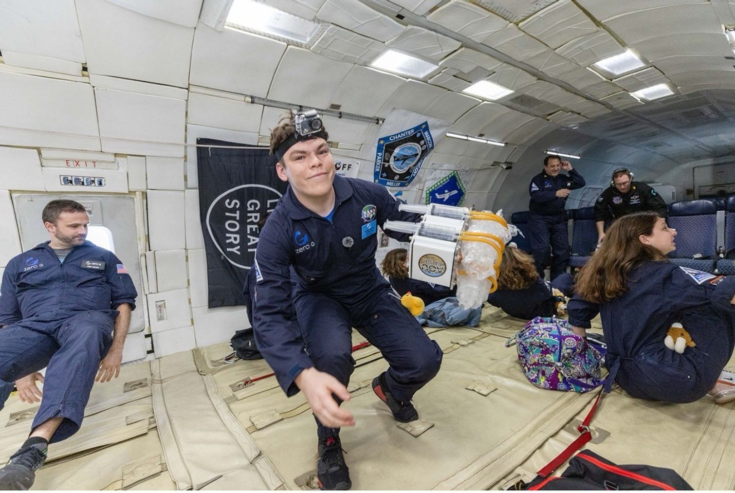 A young man in a navy blue flight suit with a head-mounted camera holds an experiment module while floating inside a Zero-G aircraft. Other participants and equipment are visible in the background.