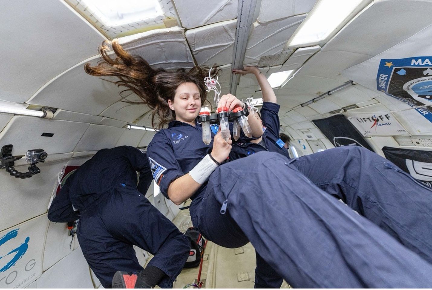A young woman with long hair floating in weightlessness examines a set of test tubes inside a Zero-G aircraft. Other participants are also floating around her.