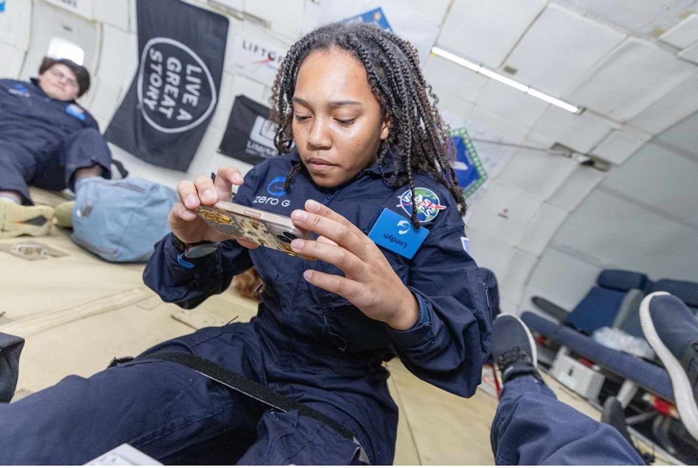 A young woman in a navy blue flight suit sits inside a Zero-G aircraft, focused on her smartphone. A teammate is reclining in the background.
