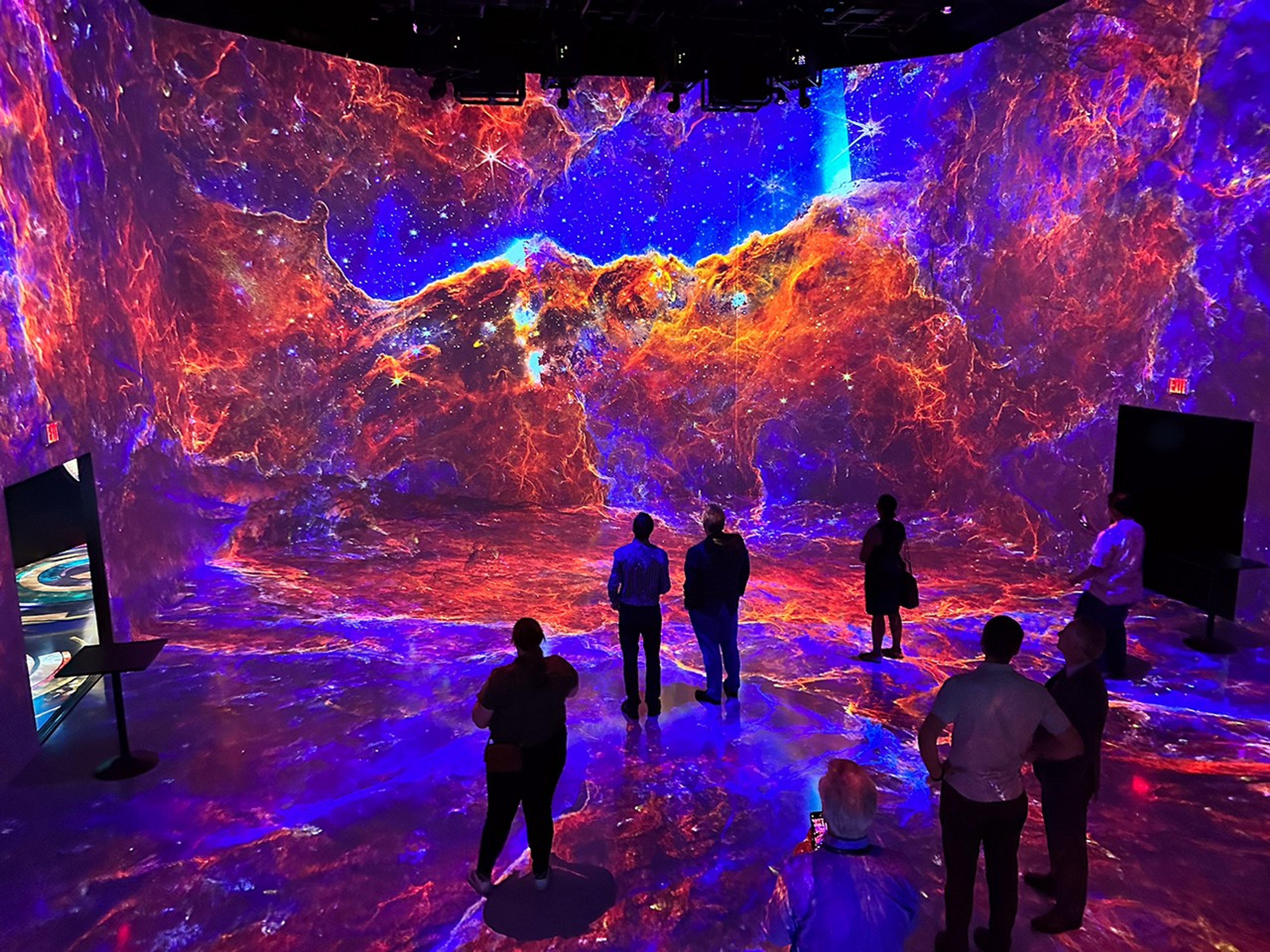 Images incorporating science data from the James Webb Space Telescope are projected onto the walls of a large room as part of a digital art piece. Seven guests stand in the room watching the show.