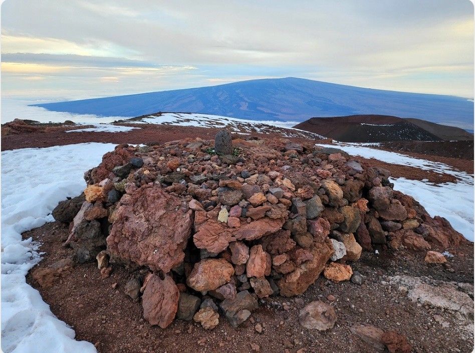 Rectangular stack of rocks on a mountain summit; another mountain is in the distant background.