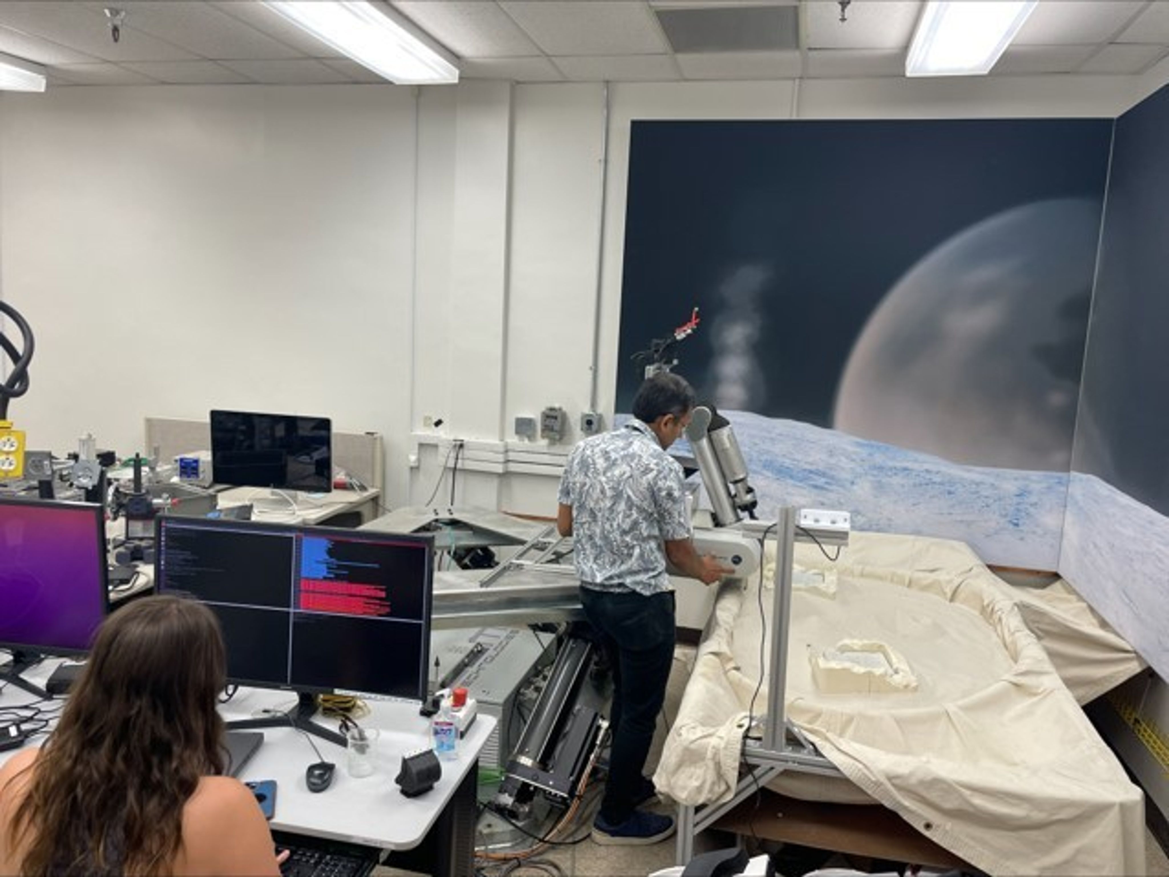 One team member at the computer station on the left side of the image is initializing the software for operating the testbed. Another team member, standing beside the robot arm, is verifying its initial configuration in preparation for use.