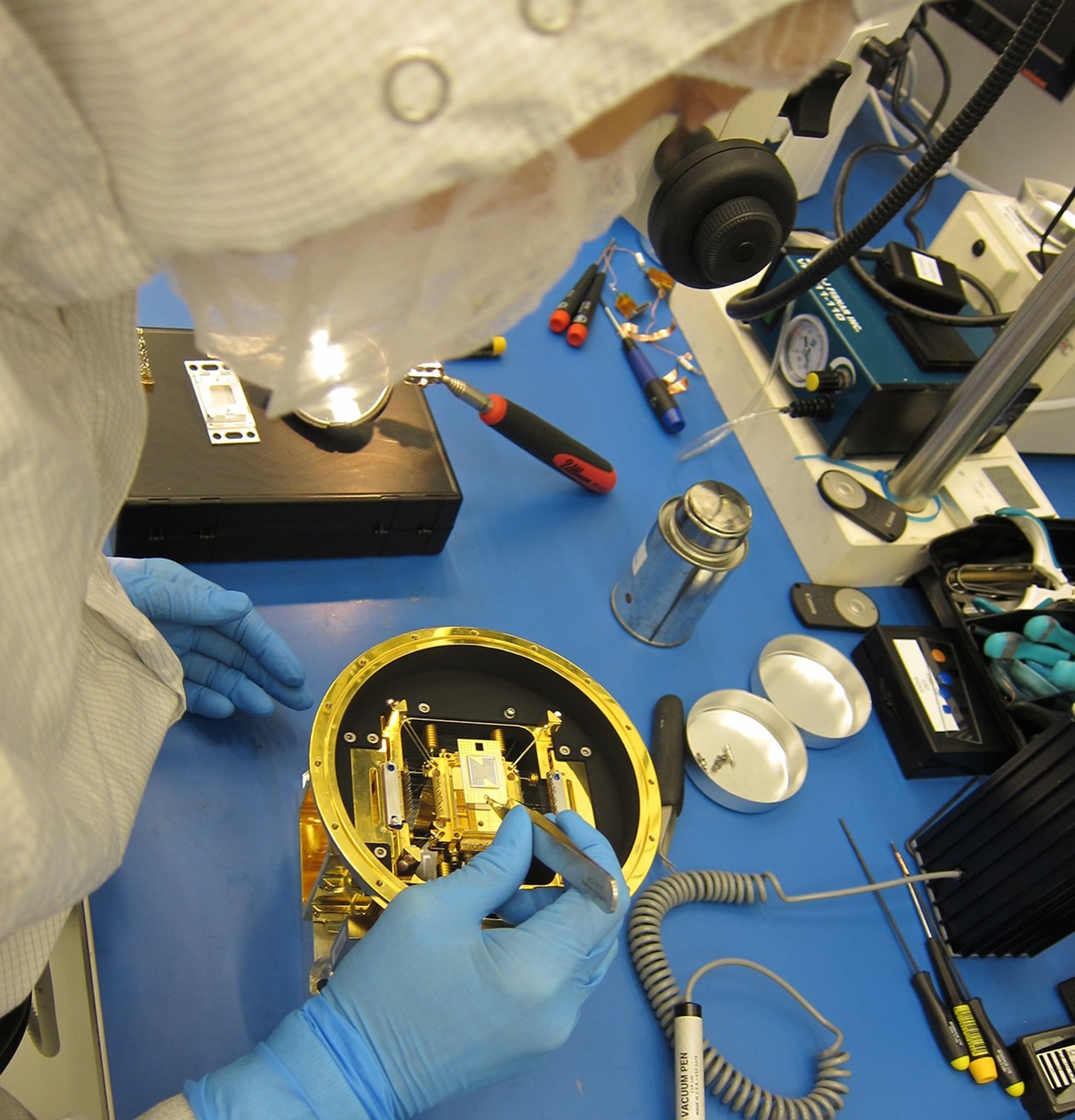 Person in white clean-room coverall, blue gloves, and face mask seated at a workbench using tweezers to place an electrical component into a gold and black assembly.