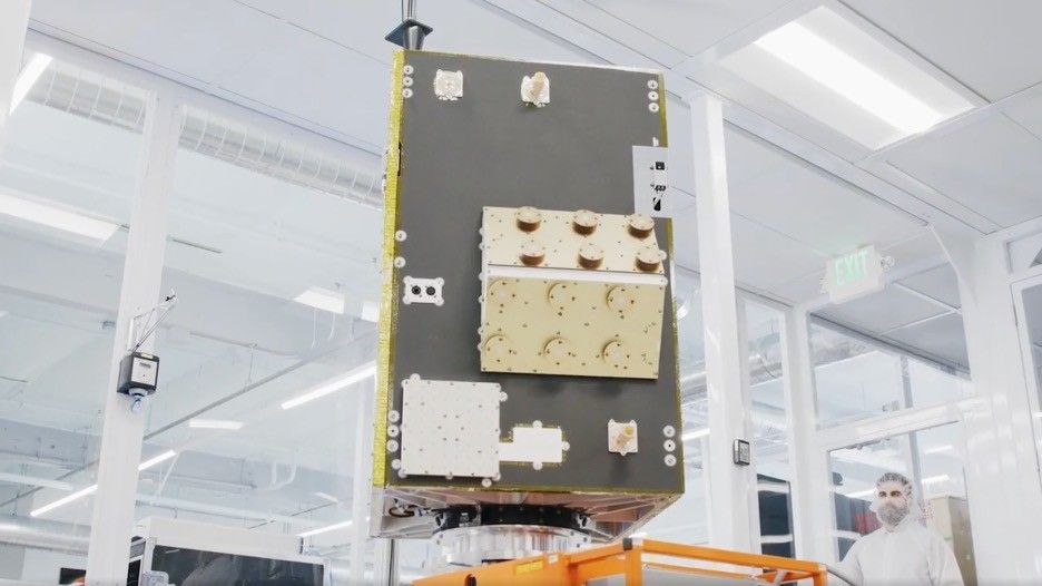 A satellite within a laboratory clean room, with a scientist in protective gear standing in the background.