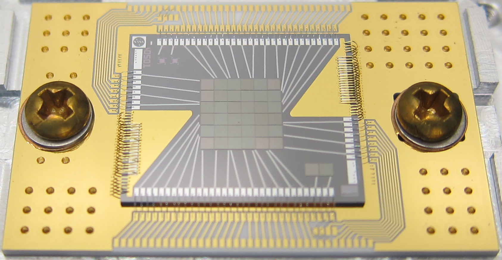 Electronic chip with wire bonds and a central square of 36 gray squares, situated atop a gold rectangle secured with screws.