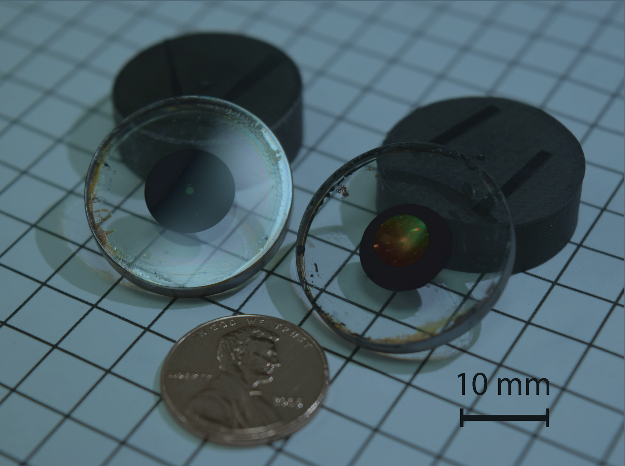 Two short black cylinders and two clear, flat, circular items with black circles in the center; all are approximately the same diameter as a penny, which is shown for scale.