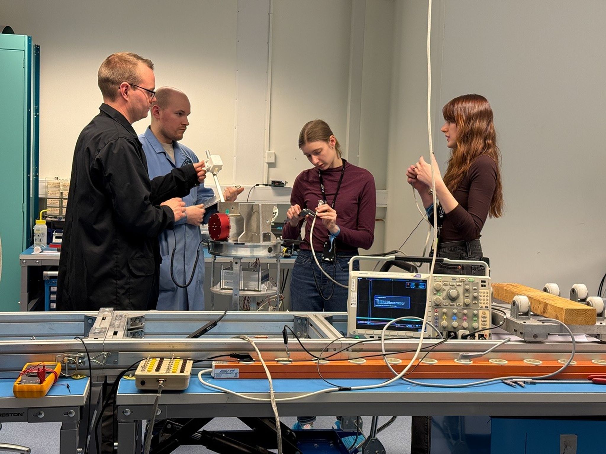 Four individuals working in a laboratory surrounded by various types of hardware on tables.